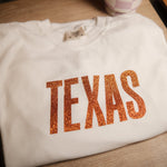 White shirt with 'TEXAS' printed in orange glitter on a wooden surface.