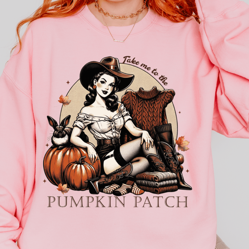 A person wearing a pink sweatshirt with a design featuring a retro cowgirl sitting with pumpkins, boots, and a hat, with the text "Take me to the Pumpkin Patch" above and below the illustration.