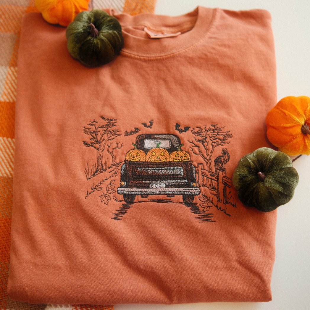 A folded orange t-shirt featuring an embroidered design of a vintage truck filled with pumpkins, driving through a fall scene with trees and bats. Styled with mini pumpkins and a cozy autumn blanket in the background.