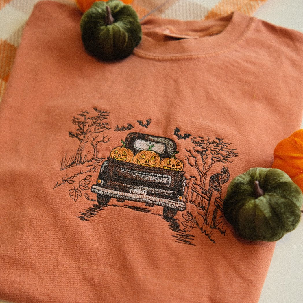 A folded orange t-shirt featuring an embroidered design of a vintage truck filled with pumpkins, driving through a fall scene with trees and bats. Styled with mini pumpkins and a cozy autumn blanket in the background.