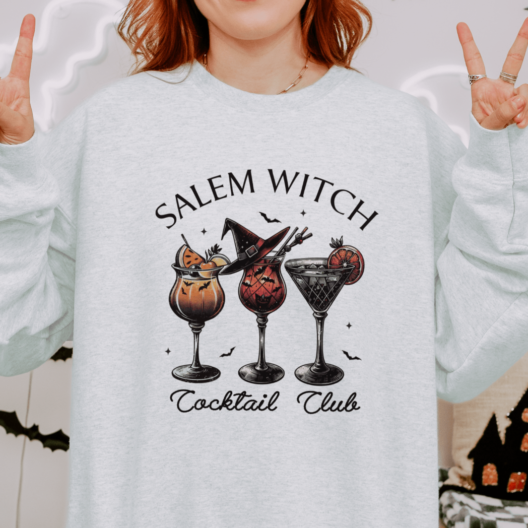 A person wearing a light grey sweatshirt with a design featuring three Halloween-themed cocktails, each with spooky decorations, under the text "Salem Witch Cocktail Club." The person is making peace signs with both hands, and the background includes Halloween-themed decorations.