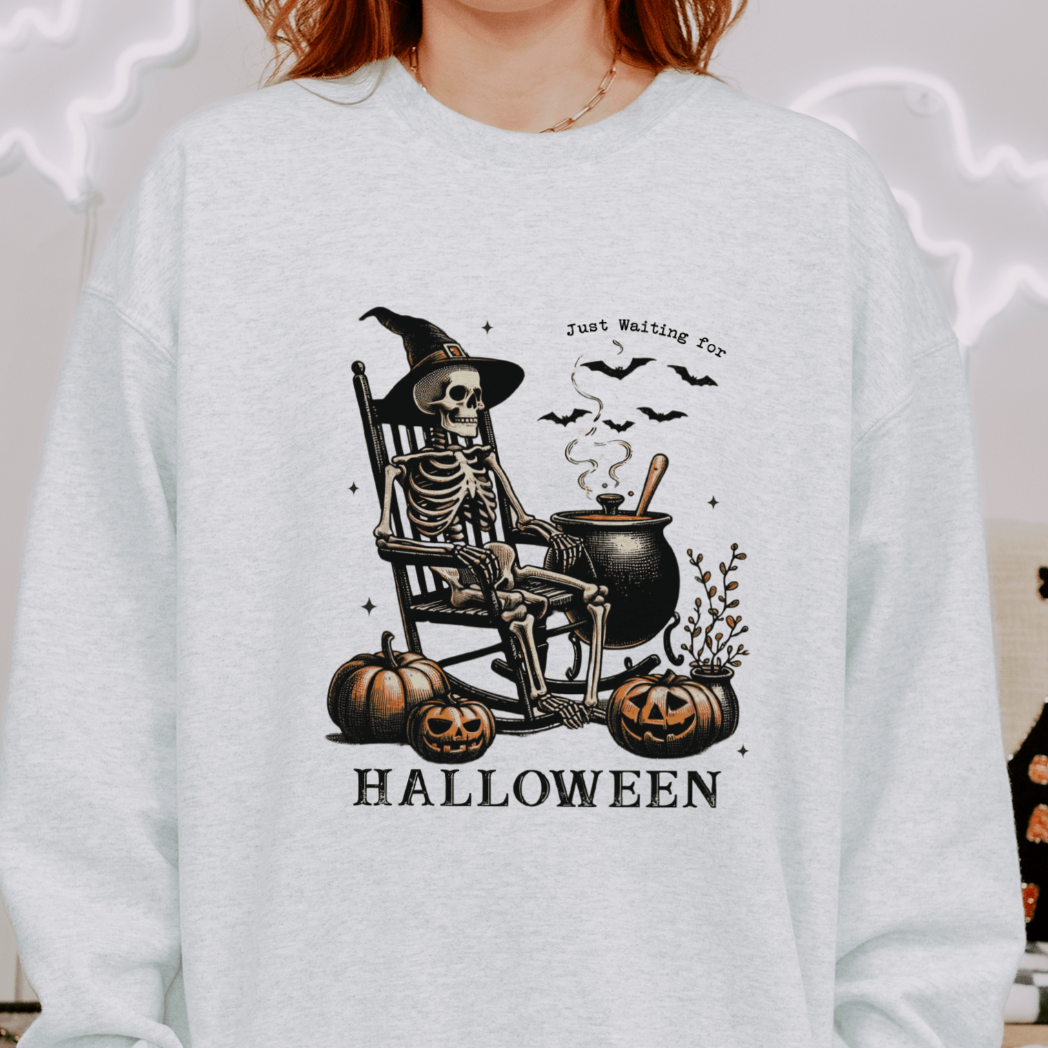 A person wearing a light grey sweatshirt with a design featuring a skeleton wearing a witch's hat, sitting in a rocking chair beside a cauldron and pumpkins, with the text "Just Waiting for Halloween" above the illustration. The background includes Halloween-themed decorations.