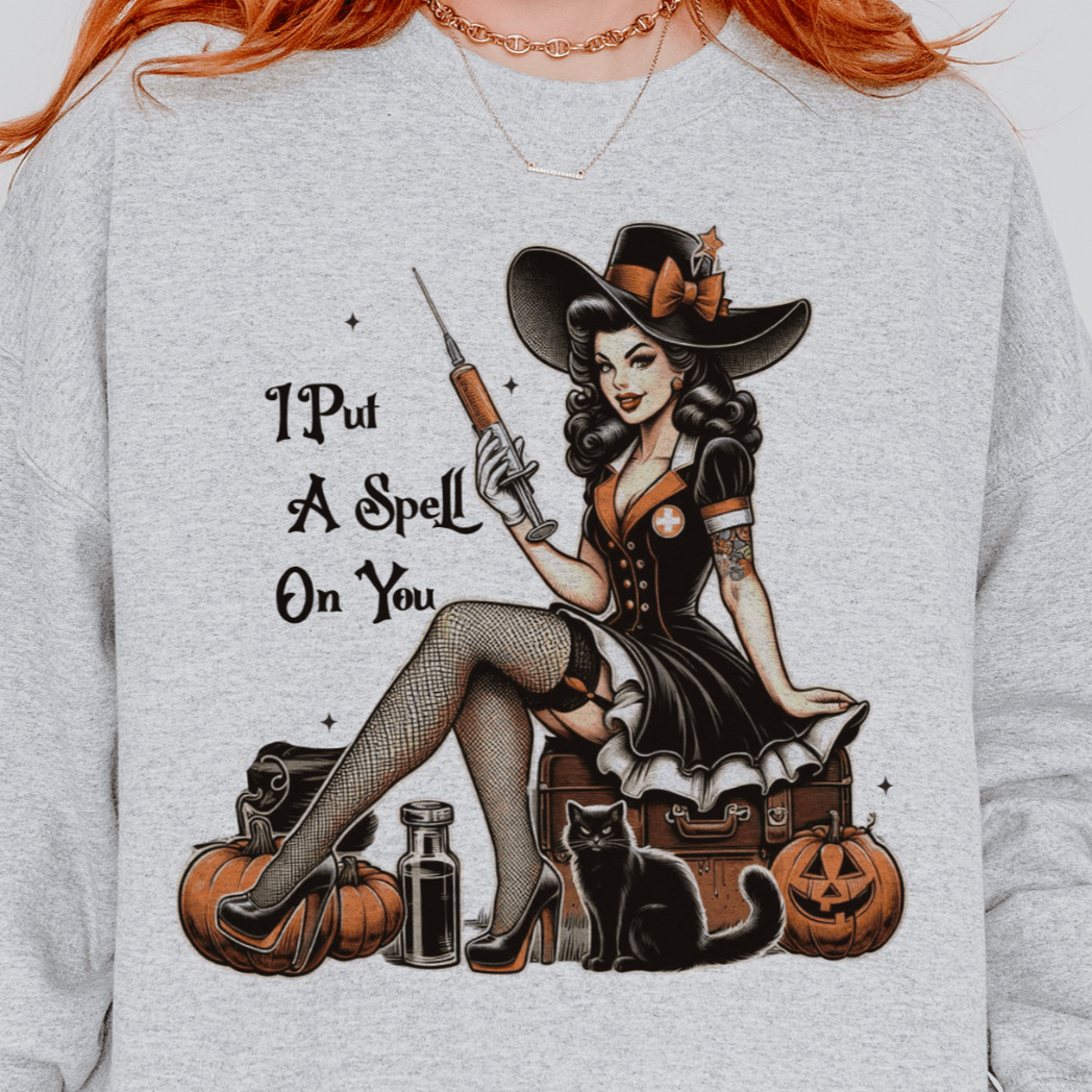 A person wearing a grey sweatshirt with a design featuring a retro witch sitting on a suitcase with pumpkins, potions, and a black cat, with the text "I Put a Spell On You" beside the illustration.