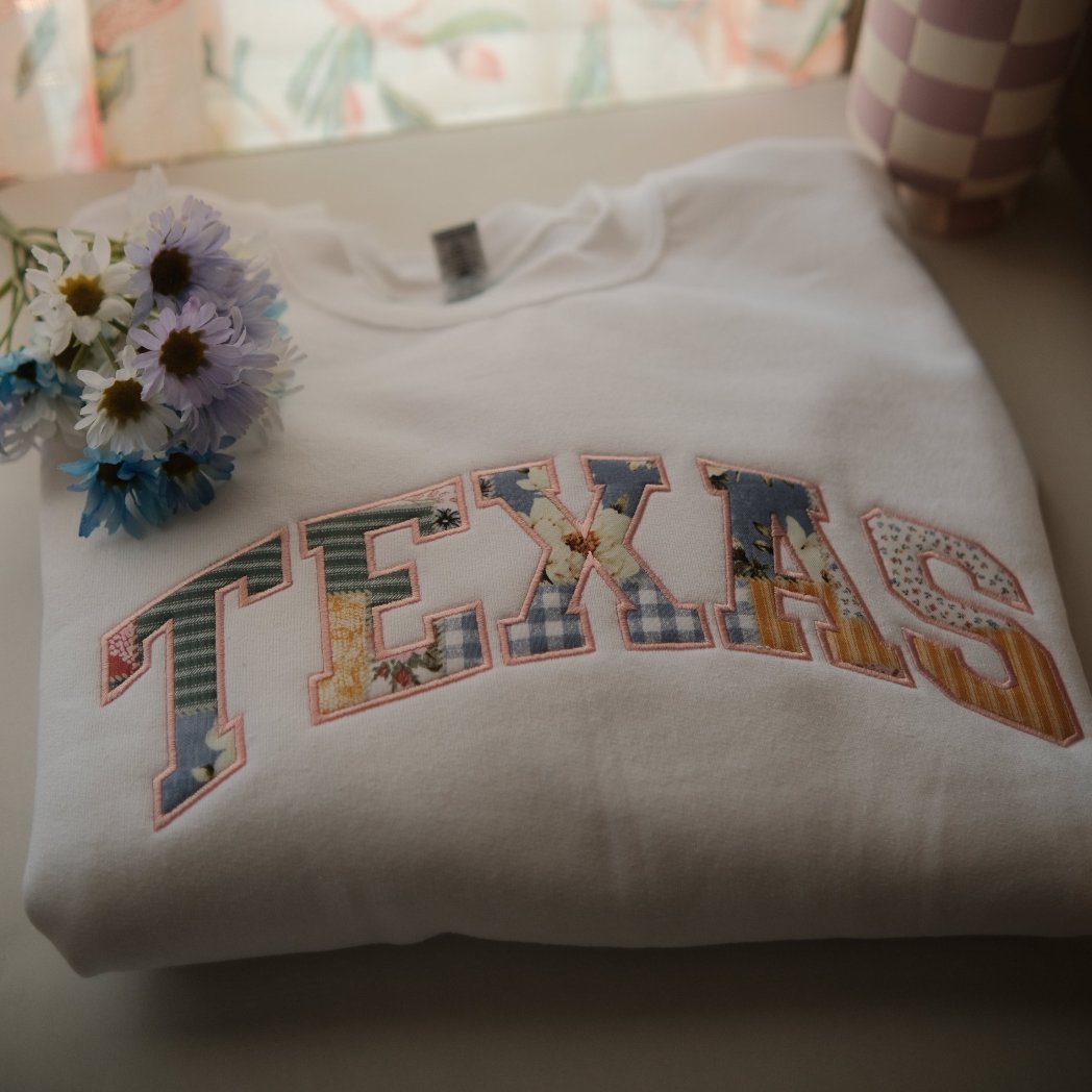 A folded white sweatshirt with the word "TEXAS" embroidered in colorful patchwork letters, featuring floral and gingham patterns. A small bouquet of flowers rests on top of the sweatshirt, adding a soft, cozy feel.