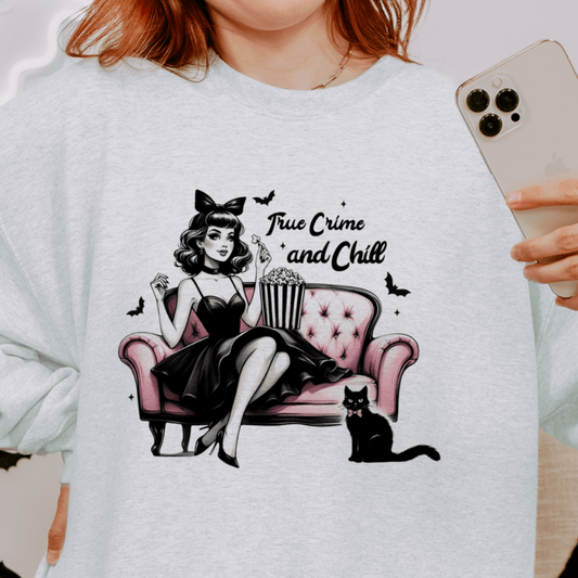 A person wearing a light grey sweatshirt with a design featuring a retro-inspired woman sitting on a pink sofa with a bowl of popcorn, accompanied by a black cat, and the text "True Crime and Chill" above the illustration. The person is holding a phone and the background includes Halloween-themed decorations.