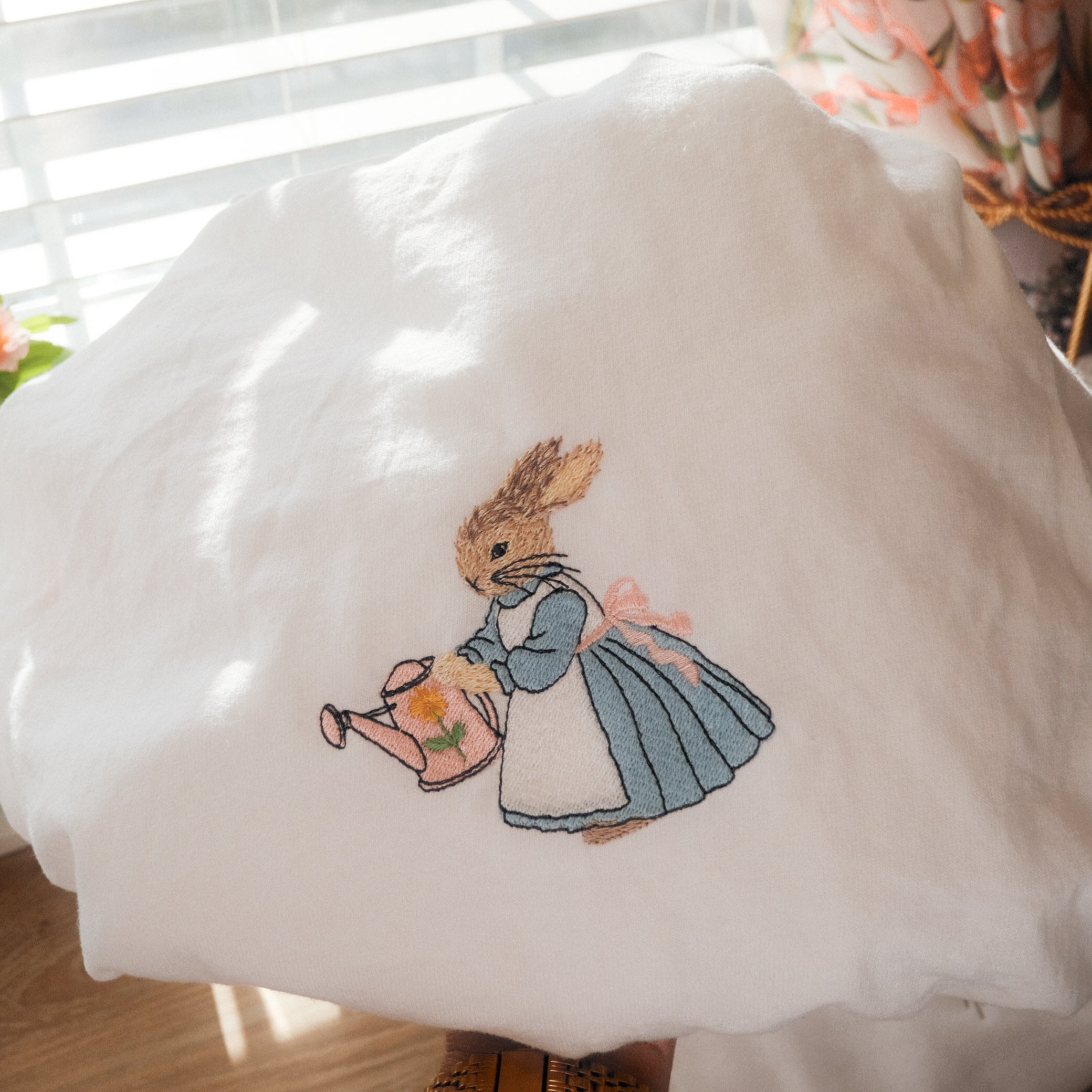 White sweatshirt with embroidered bunny wearing a blue dress and watering flowers, styled with pastel florals