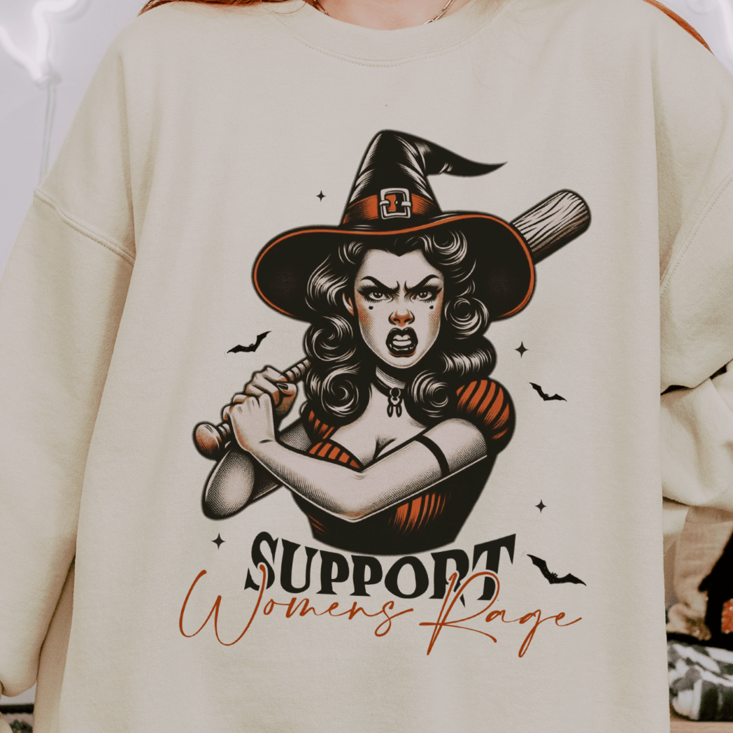 A person wearing a beige sweatshirt with a design featuring a fierce retro witch holding a bat with the text "Support Women's Rage" below the illustration. The witch is depicted with an intense expression, surrounded by small bats.