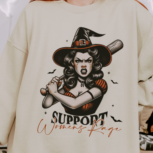 A person wearing a beige sweatshirt with a design featuring a fierce retro witch holding a bat with the text "Support Women's Rage" below the illustration. The witch is depicted with an intense expression, surrounded by small bats.